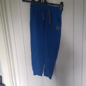 Blå mjukisbyxor från Lupilu 122/128 - Blå mjukisbyxor från Lupilu med grå snörning i midjan och ribbade muddar vid benslut. Byxorna har två fickor fram och en cool patch med motiv på ena benet. Perfekta för chill dagar eller träning.