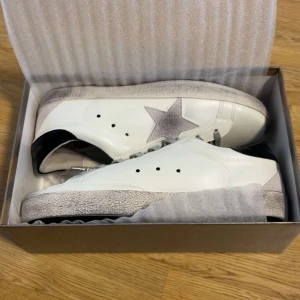 Golden Goose vita sneakers med stjärna - Snygga vita sneakers från Golden Goose med grå mockadetaljer och ikonisk stjärna på sidan. Skorna har grå snörning, svart hälkappa och slitna detaljer på sulan för en cool, urban look. Tillverkade i skinn och mocka, perfekta för dig som gillar streetstyle.