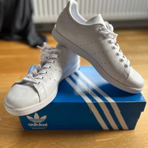 Vita Adidas Stan Smith sneakers - Klassiska helvita Adidas Stan Smith sneakers i skinn med perforerade ränder på sidorna och diskret logga på häl och plös. Snygg minimalistisk look med rund tå, snörning och platt sula. Perfekta för en clean och stilren outfit. Aldrig använda endast provade. Nypris ca 1300