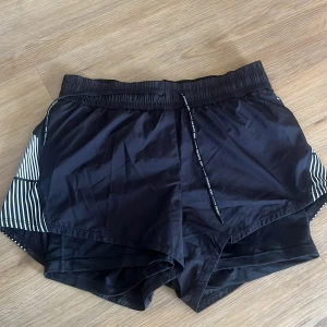 Svarta sportshorts från H&M med snörning - Svarta sportshorts från H&M Sport i polyester med vita ränder på sidorna. De har elastisk midja med snörning och en loose passform, perfekta för träning eller löpning. Snygg kontrast med de randiga detaljerna på sidorna som ger en sportig vibe. Har inbyggda tighta shorts.