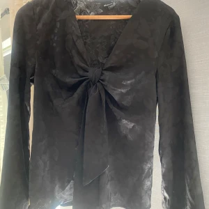 Svart blus med knyt från Gina Tricot - Supersnygg svart blus från Gina Tricot med subtilt blommönster i tyget. Djup v-ringning och stor knytrosett framtill ger en trendig look. Långa ärmar och lätt glansigt material som känns lyxigt. 