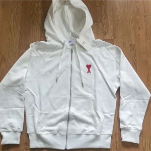 Vit hoodie med röd hjärtlogga från Ami - Vit hoodie från Ami med dragkedja, huva och snörning. På bröstet sitter en röd hjärtformad logga. Tröjan har kängurufickor och ribbade muddar. Perfekt för en chill och stilren look.