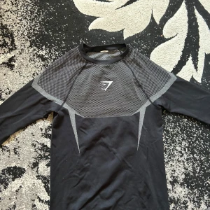 Gymshark onyx longsleeve - Svart långärmad träningströja från Gymshark med gråa detaljer och mönster över axlar och rygg. Tight passform och tillverkad i ett stretchigt syntetmaterial som andas. Perfekt för gymmet eller löprundan.