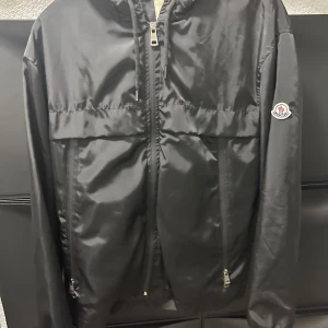 Svart vindjacka från Moncler M - Säljer en svart vindjacka från Moncler i storlek M. Jackan har huva, dragkedja framtill och två fickor med dragkedja. Materialet är glansigt och lätt, perfekt för blåsiga dagar. Moncler-logga på ärmen och snygga detaljer invändigt.