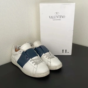 Valentino Garavani Open sneakers vit/blå - Snygga Valentino Garavani Open sneakers i vitt skinn med bred marinblå läderdetalj över snörningen. Klassisk rund tå, platt sula och diskret logga på plösen. Perfekta för dig som gillar lyxiga och stilrena sneakers med unik touch. Bra behandlade och väldigt bekväma. Priset inte hugget i sten. 