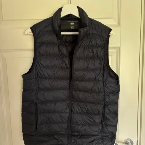 Svart dunväst från Uniqlo, S - Säljer en svart, ultralätt dunväst från Uniqlo i storlek S. Västen har quiltad design, hög krage och dragkedja framtill. Perfekt för lager-på-lager och enkel att packa ner i väskan. Stilren och funktionell för kyliga dagar.