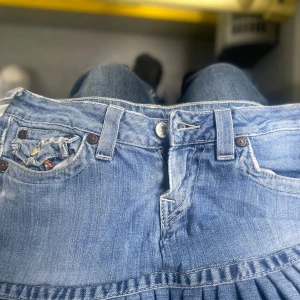 True Religion jeanskjol med brodyr - Säljer en blå jeanskjol från True Religion med plisserad nederkant och färgglada broderier på bakfickorna. Kjolen har klassiska jeansdetaljer som bälteshällor, knappar och fickor. Perfekt för dig som gillar streetstyle och unika detaljer.
