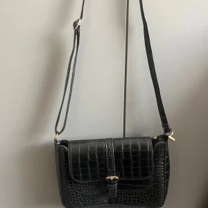 Svart axelväska i croco-imitation - Snygg svart axelväska med croco-mönstrad yta och justerbar rem. Väskan har guldfärgade metalldetaljer, spänne framtill och dragkedja under locket. Perfekt storlek för det viktigaste och ger en cool vibe till din outfit.