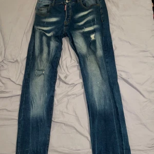 DSQUARED2 blå slitna jeans - Snygga blå jeans från DSQUARED2 med slitningar och patchade hål på benen. Klassisk femficksmodell med DSQUARED2-logga vid fickan och stor label bak. Jeansen har en rak passform och coolt tvättad look som ger en trendig vibe. (Inte äkta)