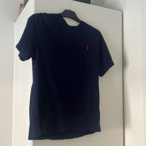 Marinblå t-shirt från Polo Ralph Lauren - Snygg marinblå t-shirt från Polo Ralph Lauren i bomull. Klassisk passform med korta ärmar och rund hals. På bröstet finns den ikoniska röda broderade polospelaren. Perfekt för en clean och stilren look.