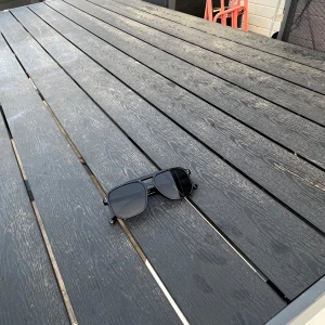 Svarta solglasögon med fyrkantig form - Snygga svarta solglasögon med breda bågar och fyrkantiga linser. Glasögonen har en modern och stilren design med mörka glas och diskreta silvriga detaljer vid gångjärnen. Perfekta för att lyfta din look under soliga dagar.