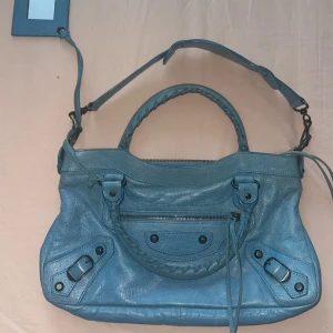Blå Balenciaga handväska i skinn - Säljer en ikonisk blå Balenciaga handväska i skinn med flätat handtag, avtagbar axelrem och spegel-detalj. Väskan har dragkedja framtill, svarta nitar och coola sömmar. Perfekt för dig som vill ha en statementväska med edgy vibe. Väskan är i jättebra stick