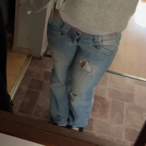 Ljusblå slitna bootcut jeans - Säljer ett par ljusblå jeans med slitningar och hål på benen. Modellen är lågmidjad med bootcut och klassiska fem fickor. Jeansen har en avslappnad vibe och är perfekta för dig som gillar en lite baggy och vintage look. Lite slitna när man gått på de