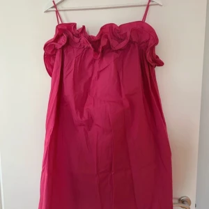 Rosa volangklänning med smala axelband - Supersöt rosa kortklänning med smala axelband och volang upptill. Lätt och luftig modell som ger en lekfull vibe, perfekt för varma dagar. Klänningen har en enkel, rak passform och är tillverkad i ett tunt bomullstyg.