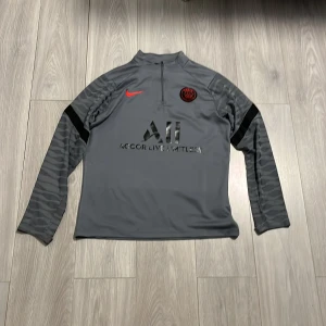 PSG långärmad träningströja Nike grå - Snygg grå långärmad fotbollströja från Paris Saint-Germain med svart och rött PSG-märke på bröstet, röd Nike-logga och svarta detaljer. Tröjan har halv dragkedja, svarta tryck på bröst och rygg samt diskreta ränder på ärmarna. Tillverkad i funktionsmaterial.