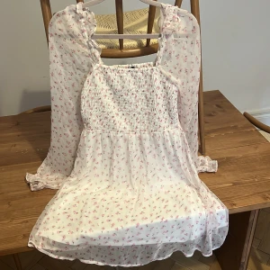 Blommig kortklänning från Vero Moda - Supersöt kortklänning från Vero Moda i vitt med små rosa blommor. Klänningen har långa, transparenta ärmar med volangdetaljer och smockad överdel. Perfekt för sommarens alla tillfällen och ger en romantisk vibe.