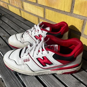 New Balance 550 vita och röda sneakers - Säljer ett par New Balance 550 sneakers i vitt med röda och svarta detaljer. Skorna har klassisk retrodesign, perforerade paneler och tydlig 550-logga på sidan. Fodret är rött och sulan har röda inslag. Perfekta för dig som gillar streetwear och sportig stil.
