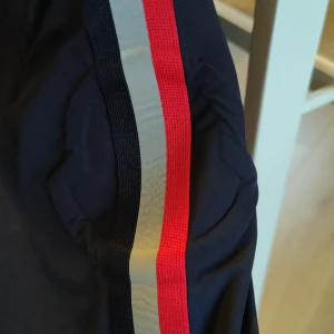 Moncler vindjacka med ränder - Mörkblå windbreaker från Moncler med huva och dragkedja framtill. Ärmarna har snygga ränder i rött, vit och blått som ger en sportig vibe. Jackan har flera fickor med dragkedja och meshfoder på insidan. Perfekt för dig som gillar stilren och exklusiv streetwear. Den är 100% äkta. Säljer billigt på grund av dens storlek. Den är storlek 130 men kan passa 140.