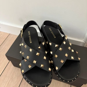 Svarta sandaler med guldnitar Sofie Schnoor - Svarta sandaler från Sofie Schnoor med breda korslagda remmar och coola guldiga nitar över hela skon. Justerbart spänne i sidan och platt sula. Tillverkade i skinn för en lyxig känsla och edgy look. Inköpta för 1200kr 