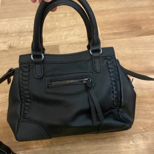 Svart handväska med dragkedjor - Snygg svart handväska i skinnimitation med både handtag och axelrem. Väskan har flera fack med dragkedjor, dekorativa sömmar och detaljer i silverfärgad metall. Perfekt storlek för att få plats med allt du behöver och passar till många olika outfits.