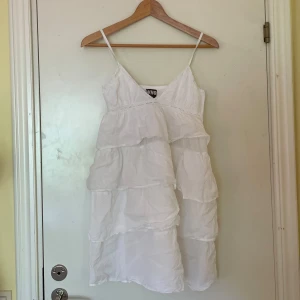 Vit volangklänning med smala axelband - Supersöt vit kortklänning från Who is she med flera lager volanger och smala axelband. Klänningen har v-ringning, spetsdetaljer och dragkedja i ryggen. Perfekt för en skolavslutning eller student!