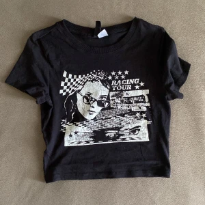 Svart croppad Racing Tour t-shirt - Svart croppad t-shirt med vit grafisk Racing Tour-print framtill. Trycket har schackrutigt mönster, stjärnor och text, samt ett par ögon och motiv i streetstil. Klassisk rund halsringning och korta ärmar. Perfekt för dig som gillar edgy och coola plagg andvänd bara 2 gr 🌸🤪
