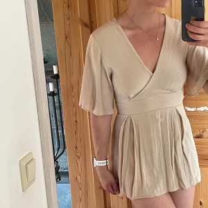 Klänning - Superfin kort beige klänning från med omlottliv, v-ringning och knyt i ryggen, har korta shorts under så man inte råkar flasha😄. Klänningen har vida korta ärmar och markerad midja. Har tyvärr växt ur den