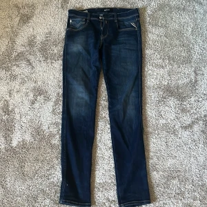 Replay Anbass mörkblå slim jeans 29/32 - Snygga mörkblå jeans från Replay, modell Anbass. Slim fit med klassisk femficksdesign och diskreta slitningar. Jeansen har raka ben och är tillverkade i stretchigt denim för extra komfort. Perfekta för dig som gillar en stilren och modern look.