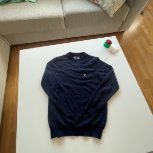 Mörkblå stickad tröja Lyle & Scott - Snygg mörkblå stickad tröja från Lyle & Scott med klassisk rund hals och gul logga broderad på bröstet. Perfekt för dig som gillar stilrena och tidlösa plagg. Långärmad modell i mjukt material som passar till det mesta.