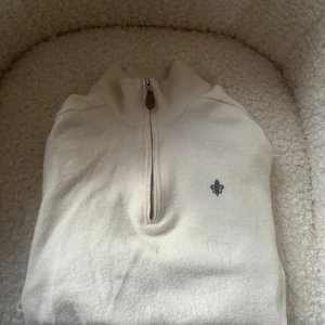 Vit merinoullströja från Morris - Snygg vit tröja från Morris i 100% merinoull med half zip och hög krage. Tröjan har en liten broderad logga på bröstet och är perfekt för dig som gillar stilrena och klassiska plagg med en modern touch.