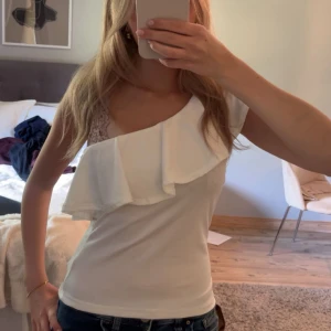Vit oneshoulder topp med volang - Säljer en vit topp med oneshoulder-design och stor volang över bysten. Toppen är figurnära och har en ärmlös sida samt en kort ärm. Perfekt för dig som gillar en stilren men ändå trendig look.