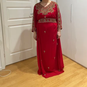 Röd långklänning med guldiga broderier /Marokansk klänning - Säljer en röd långklänning med vackra guldiga broderier och detaljer längs halsringning, ärmar och midja. Klänningen har långa ärmar och en lös passform, perfekt för dig som vill sticka ut med eleganta detaljer och lyxig känsla.