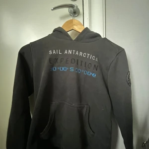 Svart hoodie Sail Racing  - Svart hoodie från Sail Racing med tryck 'Sail Antarctica Expedition' och blå detaljer på ryggen. Klassisk ficka framtill, huva med broderad logga och lång ärm. Perfekt för dig som gillar äventyr och sportig stil.