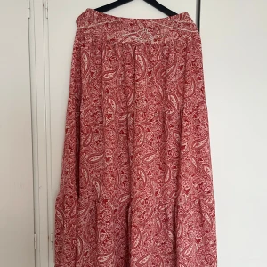 Röd långkjol med paisleymönster - Flowig röd långkjol med vitt paisleymönster och pärlbroderier upptill. Kjolen har en bohemisk vibe och är perfekt för dig som gillar färg och detaljer. Materialet känns lätt och luftigt, vilket gör den skön att bära under varmare dagar. Vissa pärlor är borta. Storlek M