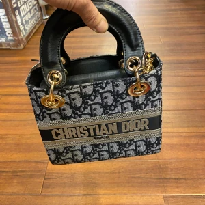  svart handväska - Snygg handväska med svart och grått monogrammönster, guldfärgade metalldetaljer och robusta handtag i svart skinn. Väskan har tydlig logga framtill och klassisk boxig form. Perfekt för dig som vill sticka ut med en ikonisk accessoar.