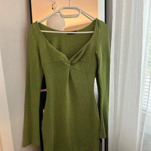 Grön långärmad kortklänning - Snygg grön kortklänning med lång ärm och twistad detalj framtill vid bysten. Klänningen har en v-ringad hals och är figurnära i modellen. Perfekt för dig som gillar en enkel men trendig look.