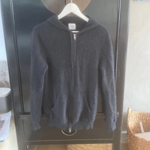 Mörkgrå cashmere hoodie John Henric - Mjuk och lyxig mörkgrå hoodie från John Henric i 100% kashmir. Tröjan har dragkedja framtill, huva och två fickor. Perfekt för dig som gillar en clean och stilren look med premiumkänsla. Passformen är normal och ärmarna är långa. Det finns små hål i högra ärmen (se bild 5) men syns knappast om inte alls när man har på sig den. Storlek M