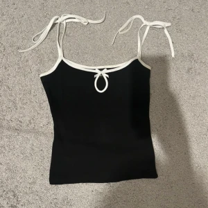 Topp från Gina Tricot - Supersöt svart topp från Gina Tricot i storlek XS. Toppen har vita kanter, smala axelband med knyt och en liten rosett framtill. Perfekt för sommaren och enkel att matcha med jeans eller kjol.