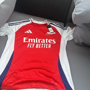 Arsenal Adidas röd matchtröja  - Arsenal fotbollströja från Adidas i rött med vita och marinblå detaljer. Klassisk rund hals, korta ärmar och ventilerande meshpaneler på sidorna. Stora loggor på bröstet och axlar med Adidas ränder. Tillverkad i lätt och snabbtorkande material.