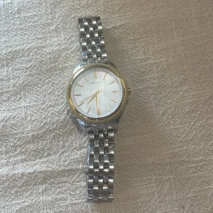 Silverfärgad klocka från Gant - Stilren klocka från Gant med länkarmband i silverfärgat rostfritt stål. Urtavlan är vit med guldiga detaljer och boetten har en guldfärgad kant. Klassisk design.