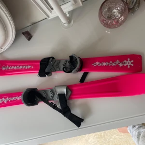 Barnskidor  -  ett par rosa barnskidor med vita snöflingemönster och svarta remmar. Perfekta för yngre barn som vill testa skidåkning på ett lekfullt sätt.