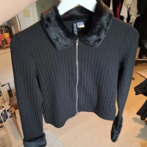 Svart ribbad kofta med fuskpäls H&M - Svart ribbad kofta från H&M med dragkedja framtill, fuskpäls på krage och ärmslut. Croppad modell med lång ärm och stilren look. Perfekt för dig som gillar trendiga detaljer och vill ha en kofta som sticker ut. Aldrig använd