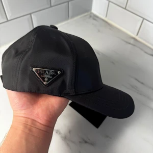 Svart keps från Prada  - Stilren svart keps från Prada med ikonisk triangellogo i metall på sidan. Kepsen är tillverkad i glansig nylon och har justerbar rem baktill för perfekt passform. En klassisk accessoar med exklusiv känsla och modern look.