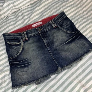 Lågmidjad jeanskjol från Stradivarius - Mörkblå, kort jeanskjol från Stradivarius med slitningar och fransig kant. Kjolen har fina veckdetaljer bak. Midjemått: ungefär 37 cm rakt över