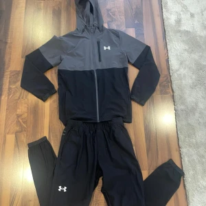 Under Armour svart och grå dress - Snygg tvåfärgad vindjacka och byxor till från Under Armour i svart och grått. Jackan har huva, dragkedja framtill och en bröstficka med zip. Perfekt för träning eller chill, med sportig look och logga på bröstet. Långärmad och tillverkad i lätt syntetmaterial.