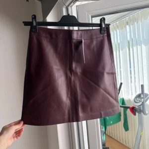 Vinröd skinnkjol i kort modell - Säljer en snygg vinröd kort kjol i skinnimitation. Kjolen har en clean och stilren design utan synliga detaljer eller fickor, perfekt för att lyfta vilken outfit som helst. Passformen är normal och materialet känns mjukt och följsamt.