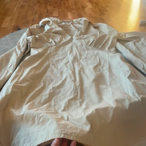 Beige skjortklänning med dragkedja från NA-KD - Snygg beige skjorta från NA-KD med markerad midja och dragkedja framtill. Skjortan har lång ärm, klassisk krage och lätt rynkade detaljer vid bysten. Perfekt för dig som gillar stilrena och trendiga plagg.storleken är 42, men passar M för de är oversize 