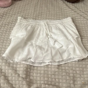 Vit volangkjol - Supersöt vit kortkjol från Gina Tricot med volanger och elastisk midja. Kjolen har dekorativa snören med tofsar och pärldetaljer framtill. Perfekt för sommarens alla tillfällen och ger en fräsch och luftig känsla. Lite defekter på snörerna men inget man tänker på. Storlek 158/164.