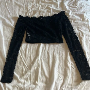 Svart croppad spets offshoulder topp - Säljer en svart croppad topp med långa ärmar och offshoulder-modell. Toppen är helt i spets med blommigt mönster och har en snygg, figurnära passform. Perfekt för dig som vill sticka ut med en elegant och trendig look.