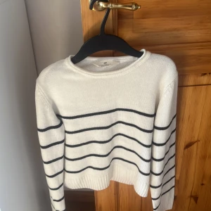 Vit och svart randig stickad tröja H&M - Vit stickad tröja från H&M med svarta horisontella ränder. Tröjan har rund halsringning, långa ärmar och ribbade muddar vid ärmslut och nederkant. Perfekt för en chill och stilren look.